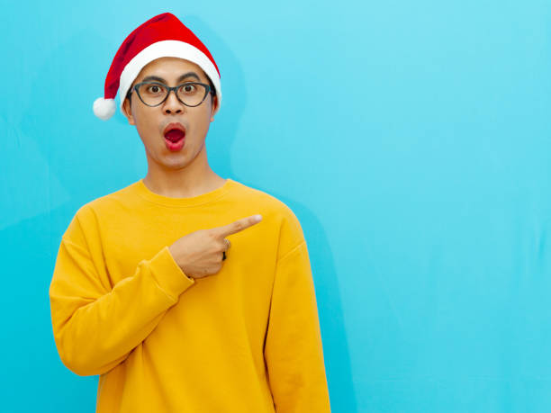 istock Asian man wearing a Christmas hat pointing to the side with a funny surprised expression. Perfect for holiday advertisements and promotions. 2177883016
