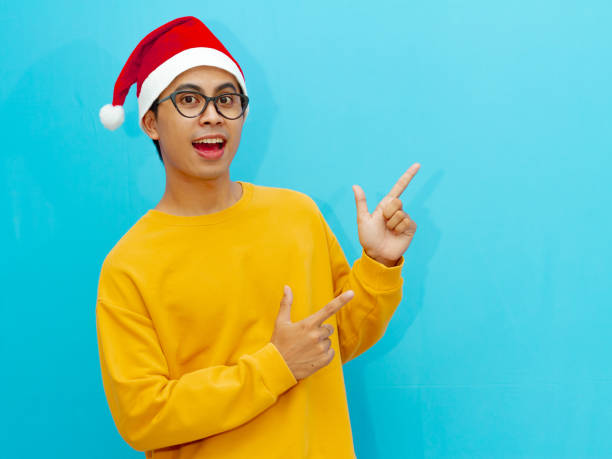 istock Asian man wearing a Christmas hat smiling happily while pointing to the side, perfect for holiday advertising or promotions. 2177883017
