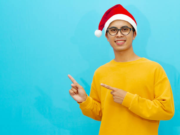 istock Asian man wearing a Christmas hat smiling happily while pointing to the side, perfect for holiday advertising or promotions. 2177883106