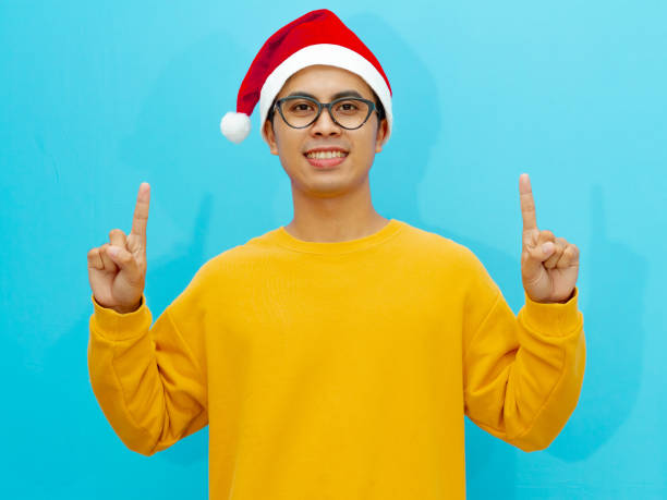 istock Happy Asian man wearing a Christmas hat, smiling and pointing up while looking at the camera, perfect for holiday advertising. 2177883108