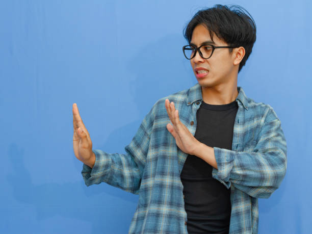 istock Serious young Asian man making a stop gesture, showing rejection or denial. Portrait on a blue background, ideal for themes of refusal, boundaries, and negative reactions. 2177884842