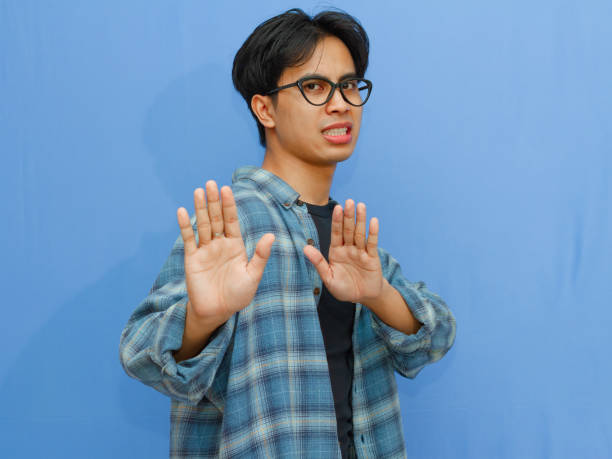 istock Serious young Asian man making a stop gesture, showing rejection or denial. Portrait on a blue background, ideal for themes of refusal, boundaries, and negative reactions. 2177884844