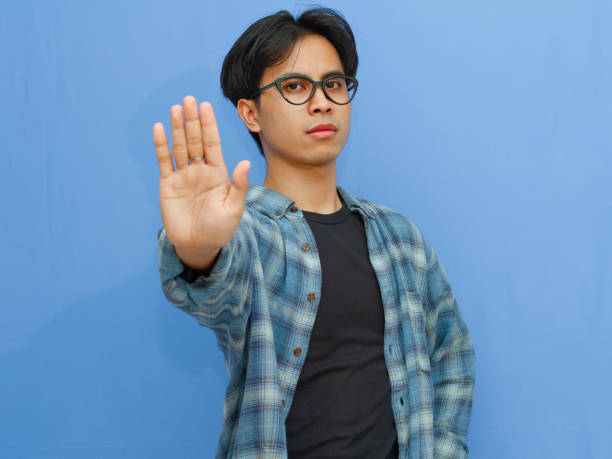 Serious young Asian man making a stop gesture, showing rejection or denial. Portrait on a blue background, ideal for themes of refusal, boundaries, and negative reactions.