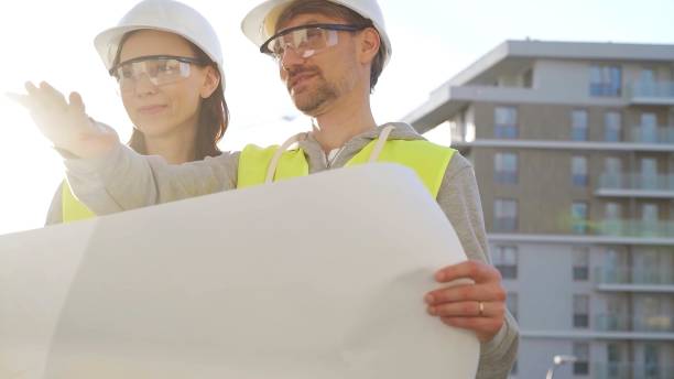 istock Two architects or engineers wearing safety hard hats and vests holding blueprint and discussing construction site project at sunrise, front view. Architecture, engineering and teamwork concept 2177906995