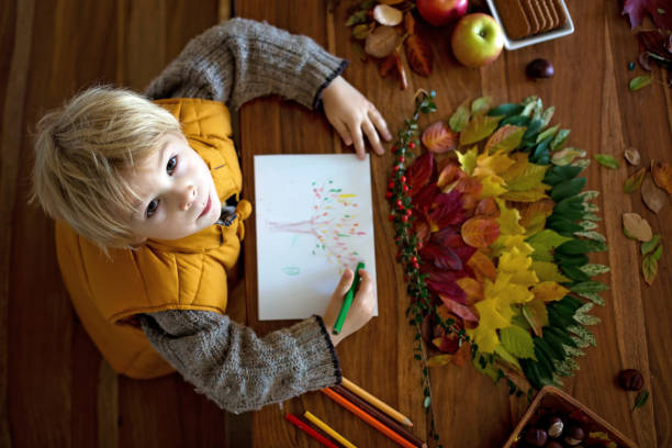 Sweet child, boy, applying leaves using glue while doing arts and chraft with leaves autumn at home, colorful spectrum of leaves