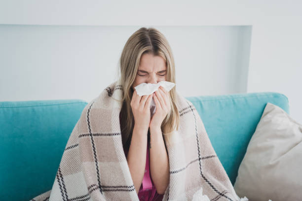 istock Young woman with cold sitting on sofa, wrapped in blanket, using tissue in living room during daytime 2177929008