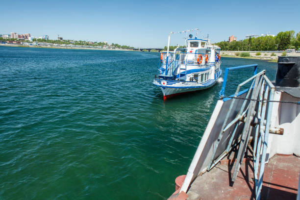 istock This photograph captures a serene river scene with a small boat or river tram moving along the calm blue water. The boat, painted white and blue, creates gentle ripples as it moves away from the camera. 2177929117