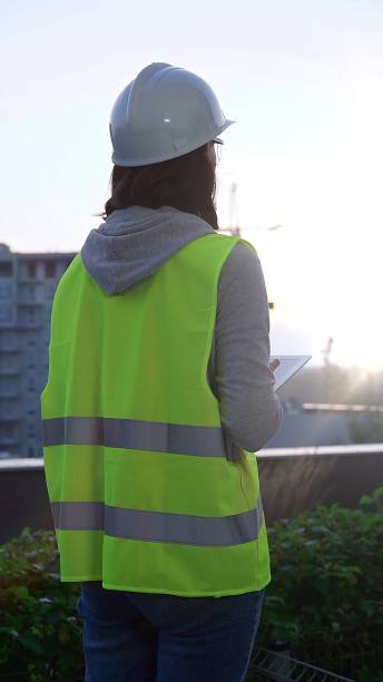 istock Woman construction engineer with a white hard hat and safety vest is using digital tablet while inspecting a construction site in the early morning at sunrise, back view 2177940668