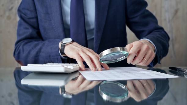 istock Auditor man analyzing financial documents with magnifying glass and calculator at his glass office desk in formal blue jacket. Business people concept 2177947069