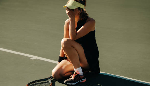 Upset woman crouched on tennis court crying after losing match A woman crouched on a tennis court, crying after losing a match, wearing a sports cap and tennis attire. athlete fails stock pictures, royalty-free photos & images