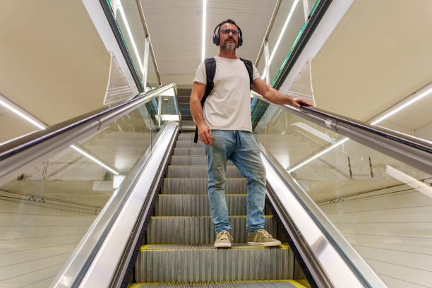 Middle-Aged Man in Casual Clothing with Backpack Descending on Escalator at Train Station to Reach Platforms