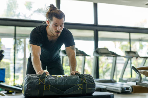 An adult man with a muscular build performs strength training by lifting a weighted bag above his shoulders. He shows dedication in his workout routine, aiming for self-improvement and fitness.
