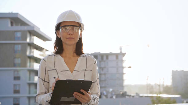 istock Woman construction engineer wearing white checked blouse and hard hat is making notes on a clipboard while inspecting a building site at sunset, front view. Architecture and engineering concepts 2177958466