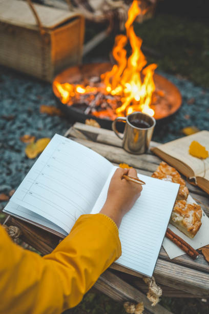istock Person writing in a notebook next to a bonfire during autumn. Outdoor cozy setting with fallen leaves, snacks, hot drink, and the warmth of the fire, capturing the essence of a peaceful autumn day 2177968494