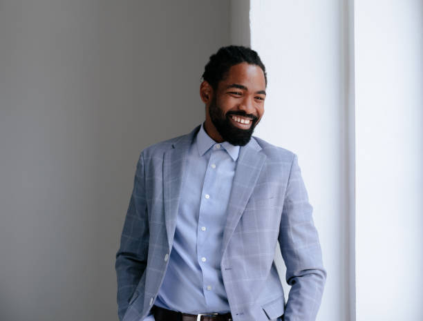 A confident businessman stands smiling near a bright window, exuding positivity and professionalism in a light checkered suit and blue shirt.