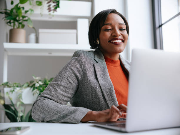 A confident professional woman works on her laptop in a modern office setting, surrounded by green plants and natural light. Her expression shows focus and positivity.