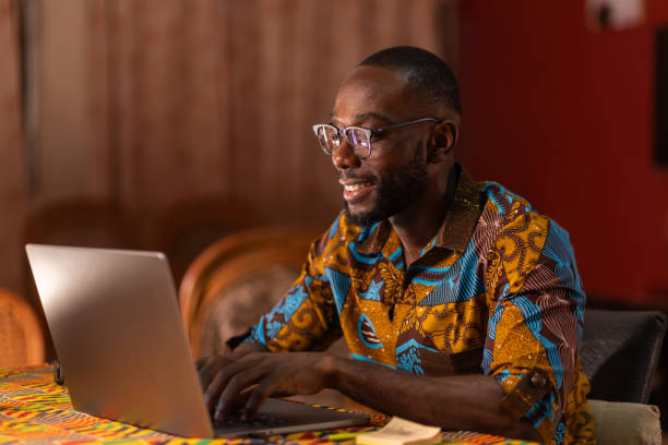 An African man wearing glasses smiles as he types on a laptop computer.