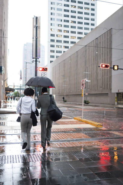 Business women walking in rainy Houston city street Two business women with umbrellas walk on a rainy city street in Houston, discussing work matters and commuting together amidst tall buildings. soaked suit stock pictures, royalty-free photos & images