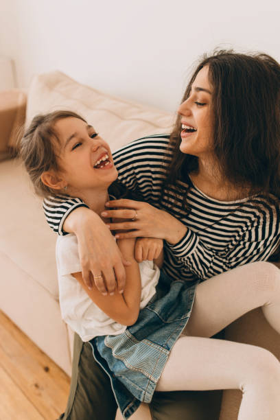 istock Vertical image of young pretty dark-haired stepmother playing with her adorable adopted daughter sitting on her knees, cuddling and laughing on couch. Human relationships. Family bonding 2177997948