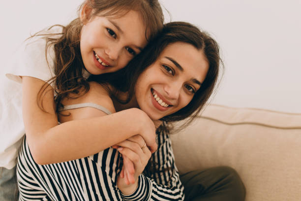 istock Little elementary school girl in white t-shirt leaning on her mother's back returned from work, hugging her, both looking happy and cheerful. Mother and daughter relationships. Family bonding 2177997968