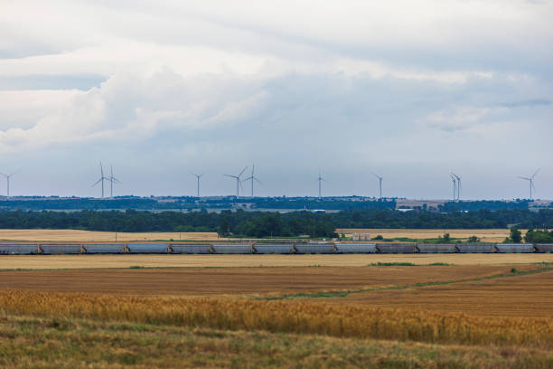 Freight train traverses golden wheat fields in rural Oklahoma. Wind farm with numerous turbines dots the horizon. Dramatic cumulonimbus clouds fill the sky, indicating potential stormy weather. Scene captures intersection of traditional agriculture, modern transportation, and renewable energy in American heartland.