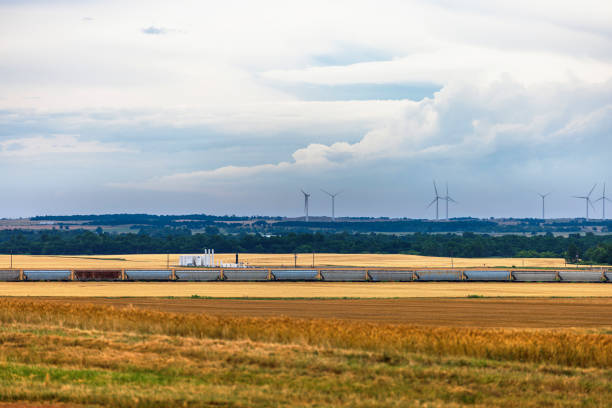 Long freight train stretches across golden wheat fields in rural Oklahoma. Vast agricultural plains dominate the landscape, with distant patches of trees. Horizon features cluster of tall wind turbines. Gathering cumulus clouds create dynamic, textured atmosphere. Scene captures intersection of traditional farming, modern renewable energy, and changing weather patterns of Great Plains