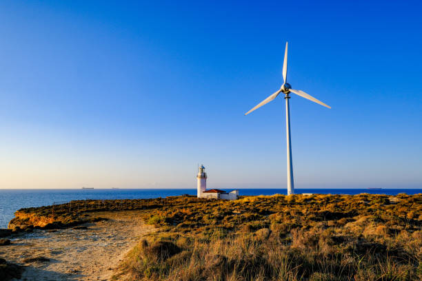 A wide-angle shot capturing the stunning scene of a lighthouse with wind turbines beside it and ships in the distance, set against a Mediterranean sunset with a clear blue sky. The lighthouse and turbines are illuminated by the warm hues of the setting sun, blending natural and modern elements into a peaceful atmosphere. Ideal for showcasing the themes of renewable energy and maritime life in the Mediterranean region.