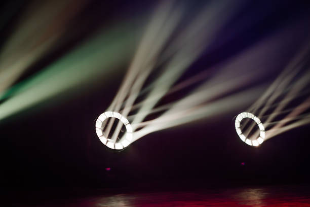 Circular stage lights emit focused beams in a dark venue, creating a dramatic effect with rays cutting through the darkness for a moody ambiance.