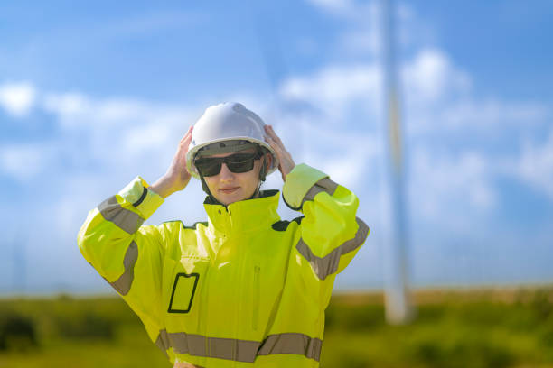 A male engineer in a checkered shirt, orange safety vest, and white helmet stands confidently on a hill at a wind power plant, with modern wind turbines in the background. The clear blue sky and vibrant greenery underline the commitment to sustainable energy solutions. The image symbolizes innovation, clean technology, and the critical role of professionals in advancing the renewable energy sector. Artificial intelligence and digital monitoring systems are becoming essential tools in managing such large-scale operations efficiently and safely