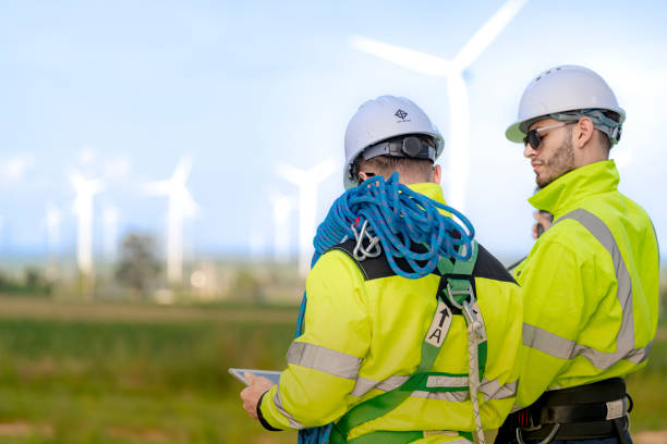 istock Two engineers in high visibility jackets and helmets are inspecting a wind farm. One is holding a mobile device, while the other is equipped with ropes for climbing, with turbines visible in the background. 2178015344