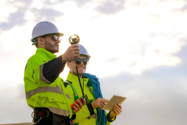 istock Two engineers wearing high visibility safety jackets and helmets measure wind speed with an anemometer and analyze data on a tablet at a renewable energy site. The cloudy sky adds depth to the scene. 2178016349