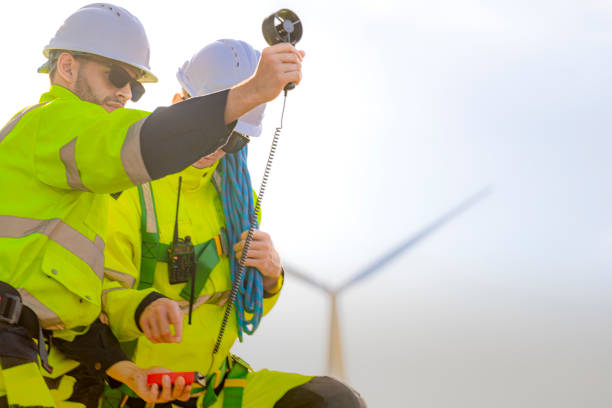 istock Two engineers wearing high visibility safety jackets and helmets measure wind speed with an anemometer and analyze data on a tablet at a renewable energy site. The cloudy sky adds depth to the scene. 2178016404