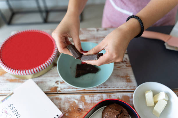 Close-up of hands preparing chocolate for holiday baking, featuring colorful tins and kitchen ingredients. A festive atmosphere with a reindeer-themed tin on a wooden table.