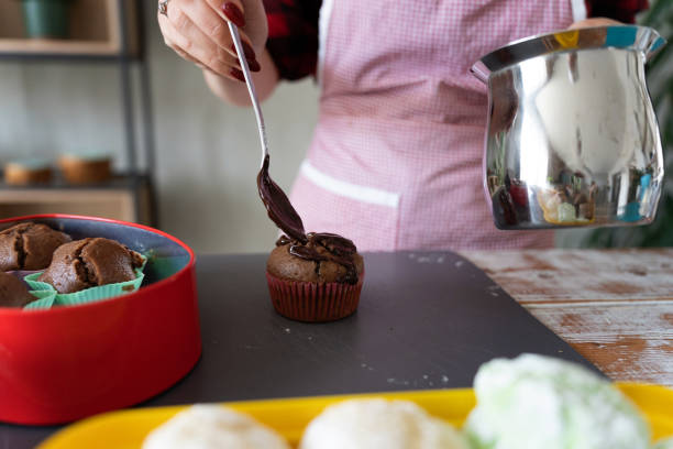 A woman carefully decorates chocolate muffins with icing in a warm kitchen environment, emphasizing the joy and art of home baking. The image highlights cozy and inviting kitchen activities.
