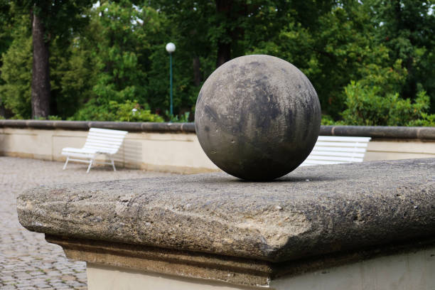 A weathered stone sphere rests on a pedestal in the garden of Prague Castle. The minimalist sculpture is set against a backdrop of greenery, cobblestone paths, and peaceful benches, creating a serene outdoor setting.