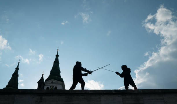 Two fencers dressed in black protective uniforms, helmets with face masks are fencing on castle wall using long medieval metal historical swords practicing before competition s. Active people concept