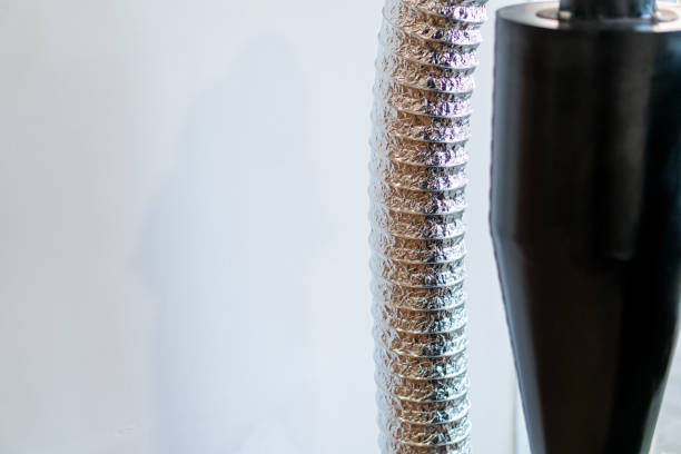 A close-up view of a metallic flexible ducting hose next to a black cylindrical object. The hose is coiled and shiny, reflecting light, while the background is a plain li