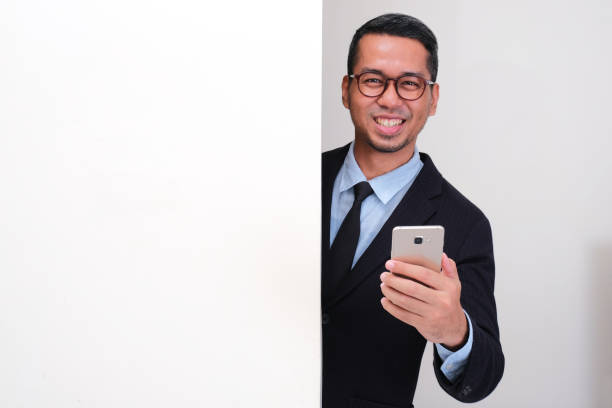 istock A businessman peeking behind wall holding mobile phone with happy face 2178057541