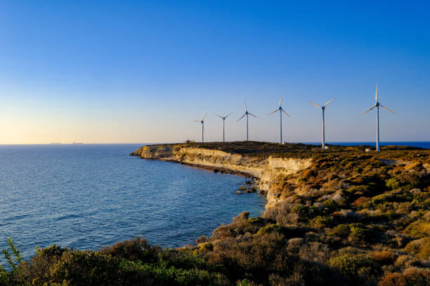 A wide-angle shot capturing the stunning view of numerous wind turbines situated on a cape at sunset in the Mediterranean. The turbines blend harmoniously with the sea view, reflecting the warm tones of the setting sun, creating a serene yet dynamic atmosphere. Ideal for showcasing renewable energy efforts in the Mediterranean region, highlighting the harmony between nature and modern technology.
