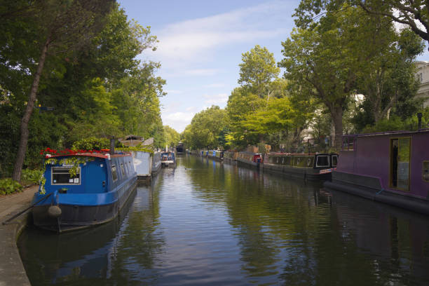 Regents Canal, London - 08 25 2024 : A view of Regent's Canal near Little Venice, London, on a sunny summer day. Colorful canal boats line the calm, tree-shaded waterway, with reflections shimmering beneath a clear blue sky.