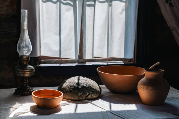 istock Rustic interior with a window bathed in sunlight. The room is dimly lit, but the window lets in a bright beam of light, highlighting a table set with a clay jug, a bowl, and a loaf of bread. 2178082881