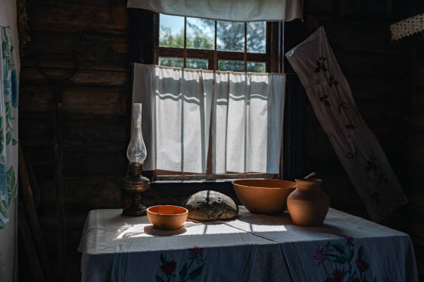 istock Rustic interior with a window bathed in sunlight. The room is dimly lit, but the window lets in a bright beam of light, highlighting a table set with a clay jug, a bowl, and a loaf of bread. 2178082895