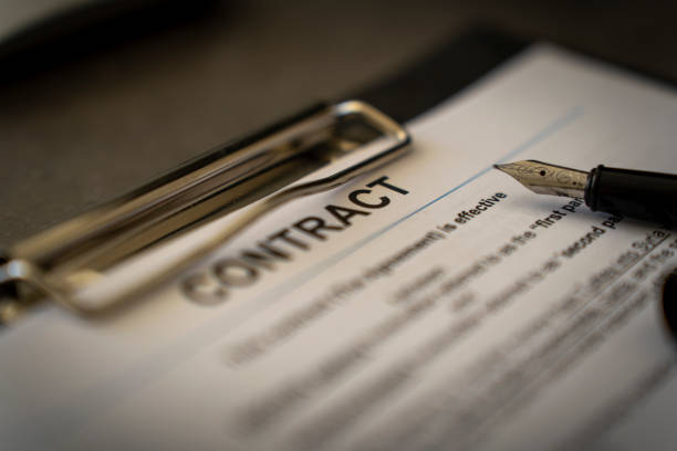A macro close up blue toned image of a fountain pen resting on sheets of legal contract paperwork documents. Show with shallow depth of field with focus on the word ‘Contract’ document title. All contact text is generic and fake.
