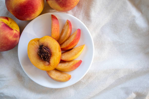Fresh peaches from the orchard, both whole and sliced for immediate consumption, are on a white plate. Peaches are a sweet and tangy fruit rich in antioxidants that help nourish the skin, making it smoother, softer, and more hydrated.
