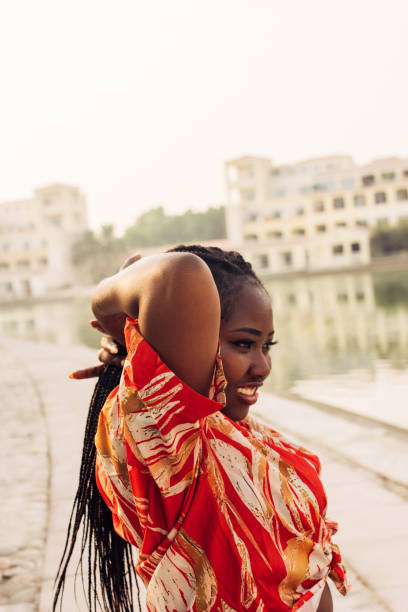 In this captivating image, a confident woman stands by the water, her expression radiant with a sense of inner peace and joy. Dressed in a vibrant, patterned outfit, she stretches her arms behind her head, basking in the warmth of the moment. The soft light reflects on the calm water, blending perfectly with the serene backdrop of the buildings in the distance. It’s a scene that captures confidence, relaxation, and the beauty of being in the present.
