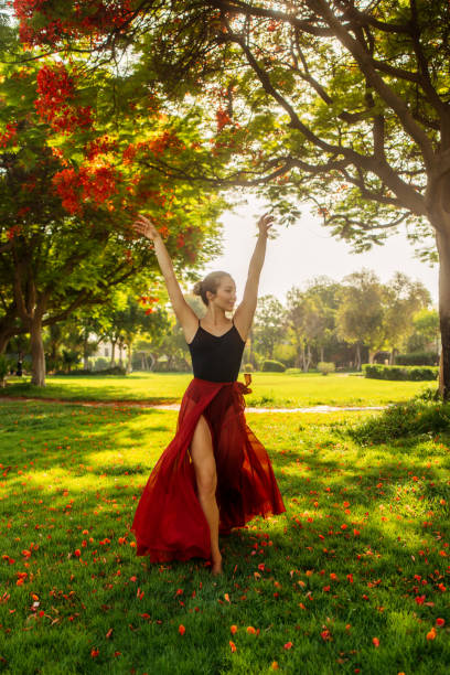 A woman gracefully dances in a sunlit garden, her red skirt billowing behind her as she moves. The vibrant greenery and sunlight cast a serene atmosphere, highlighting her lightness and joy. Dressed in a flowing red skirt and a black top, she embodies freedom, expression, and elegance, creating a captivating moment of movement and beauty. The warm sunlight filtering through the trees enhances the natural, outdoor setting.