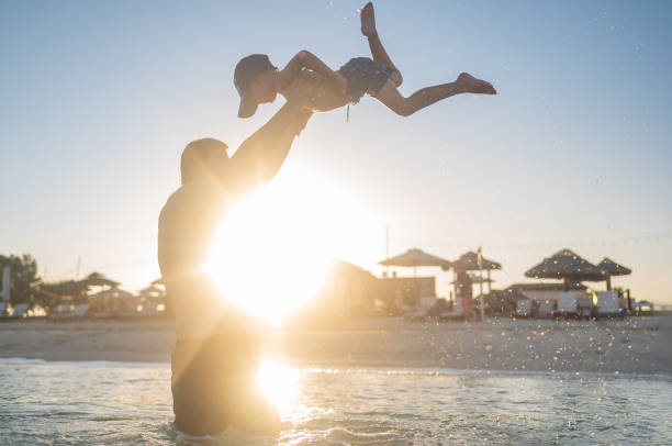 Silhouetted father joyfully lifting his child in the air on a serene beach as the sun sets, depicting family fun and bonding.