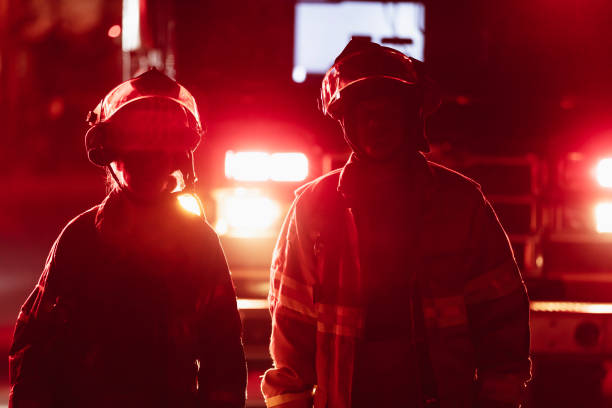 Silhouette of two multiracial firefighters wearing helmets and fire protection suits working at night. They are in front of a fire engine with headlights on, walking side by side. The female firefighter is a mid adult Filipino woman in her 30s. Her coworker is a mature Hispanic man in his 40s. They are looking toward the camera.