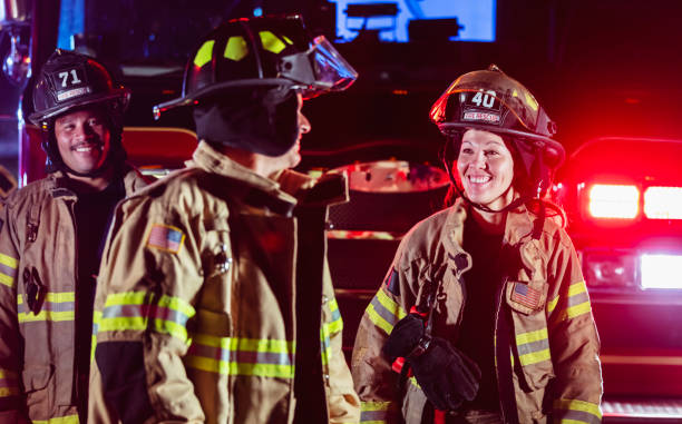 A multiracial group of three firefighters working at night wearing helmets and fire protection suits, smiling. The focus is on the female firefighter, a Filipino woman in her 30s, who is looking toward her Hispanic coworker. They are in front of a firetruck with headlights illuminated behind them.
