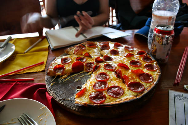 A pepperoni pizza sitting on a rustic restaurant table while someone writes their travel diary with a slice missing. There's bottles of water and soda nearby and serviettes